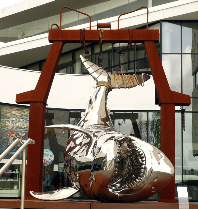 Un porche en métal avec des palans desquels pend une sculpture de requin chromé dont la gueule posée sur le sol est ouverte et montre ses nombreuses dents. L'effet est très agressif