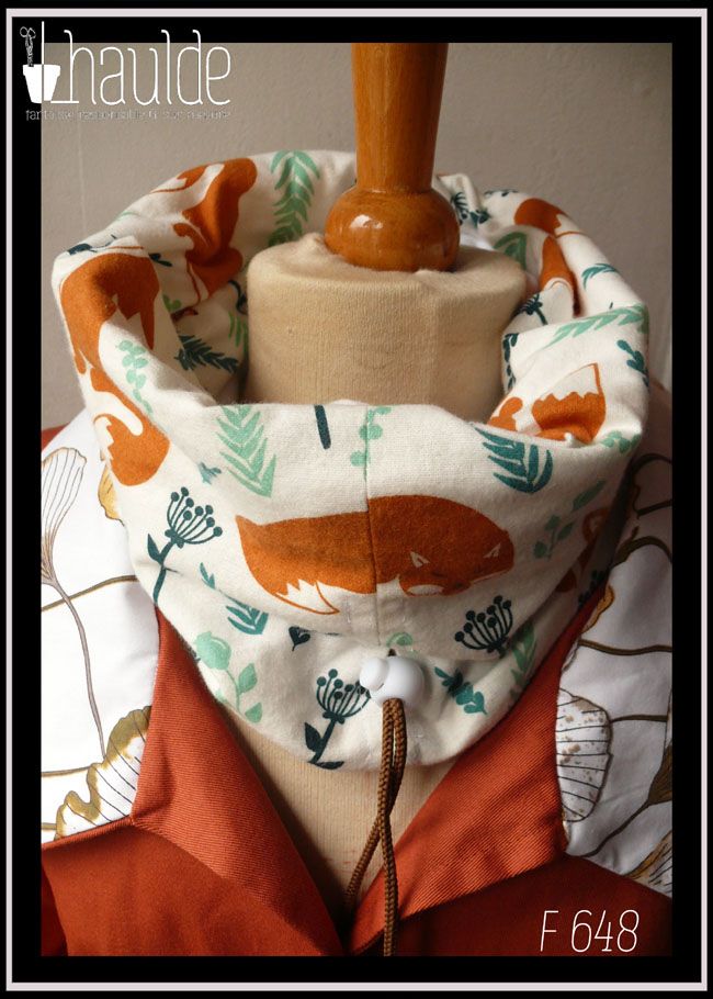 Tour de cou en tissu blanc imprimé renard roux et végétaux clairsemés dans les tons verts, le dessin est de style naïf, les renards sont dans plusieurs postures, devant il apparait couché, mais il y en a aussi des assis Cordon de serrage marron avec un stop blanc Vu sur un mannequin de couture