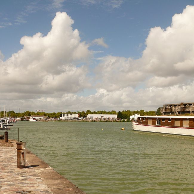 un quai au bord de l'eau couverte de vaguelettes, une péniche amarrée en face et en fond d'autres quais et des arbres sous le ciel bleu nuageux. Il s'agit des bassins à flot à Bordeaux
