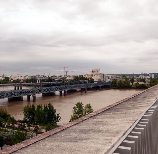 Vue d'en haut de l'ancien pont ferré sur la Garonne et de la rive droite, avec des immeubles et des grues et quelques arbres. A l'horizon les collines de la banlieue, au premier plan le parapet de la méca. Le ciel est gris.