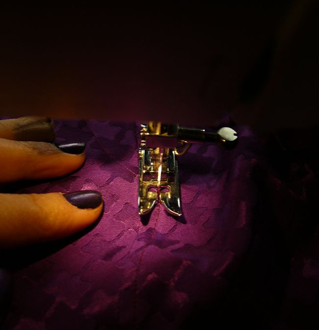 gros plan du pied et de l'aiguille de ma machine en train de piquer un tissu violet jacquard à motifs presque pied de poule, le tissu est légèrement brillant, mes doigts posés dessus portent du vernis violet