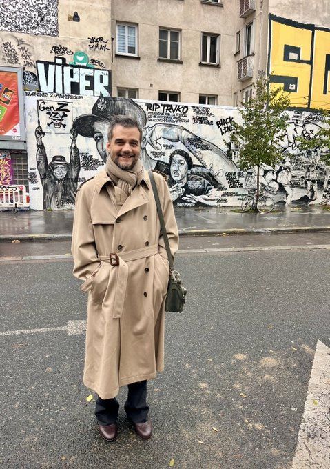Wagner Moura, em uma rua de Paris, de frente pra foto, sorrindo, com uma calça preta, sapatos marrons, um casaco bege, assim como a echarpe ao redor do pescoço, uma bolsinha pedurada e as mãos no bolso do casaco. Ao fundo, uma parece branca com desenhos em preto e branco.