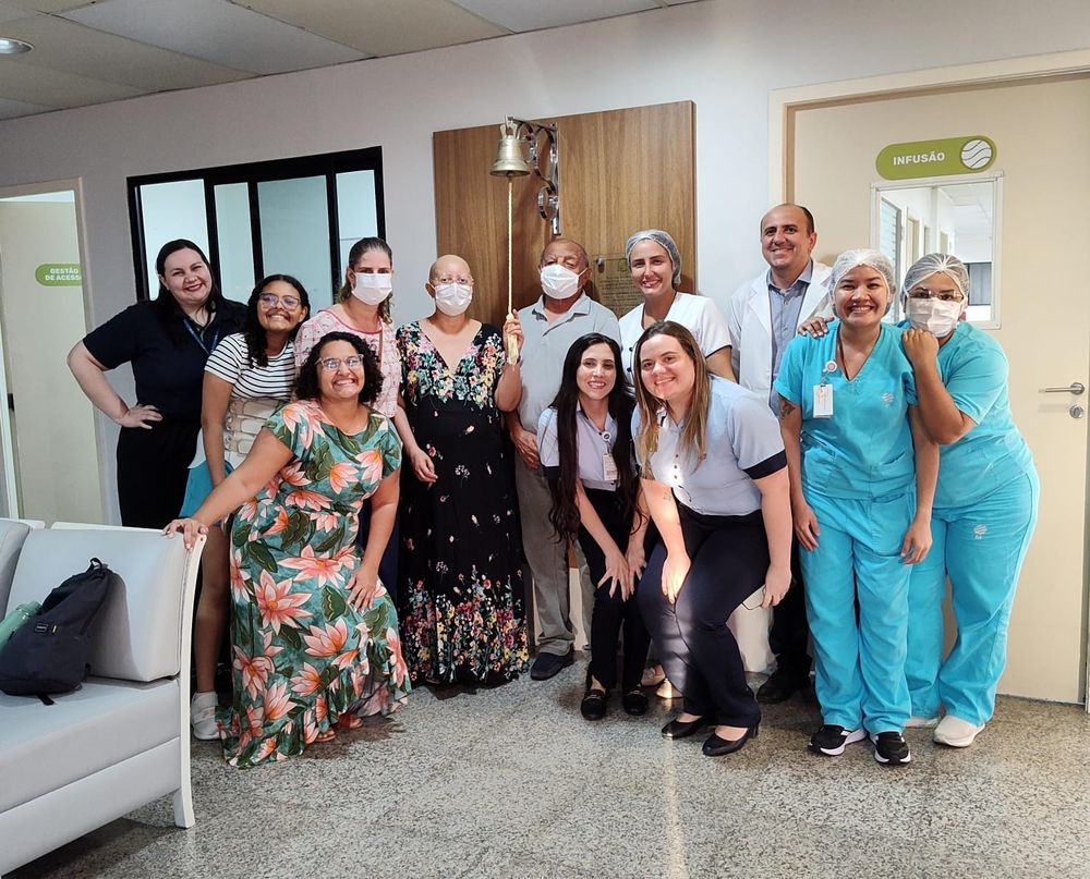 Foto com 12 pessoas encarando a câmera, 2 técnicas de enfermagem, 2 enfermeiras, 2 atendentes, meu pai, minha irmã, minha cunhada, minha sobrinha e eu, tocando o sino que indica o fim do tratamento de quimioterapia 