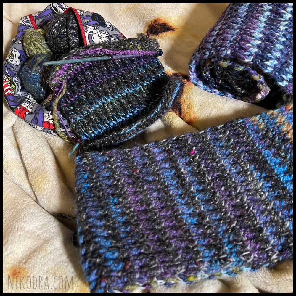 Photo close up showing parts of a tunisian crochet project that currently look like scarves of mostly handspun yarns. The one in front is folded up showing a long section of the colors. One in the upper right corner is rolled up. The last one in the upper left corner is sitting on top of a project bag that is unzipped & showing some of the yarn balls inside. This is the current WIP with a crochet hook in it that is just the start of a strip & currently looks like a square. 
The colors of all these are varieties of blues, purples, greens & black.