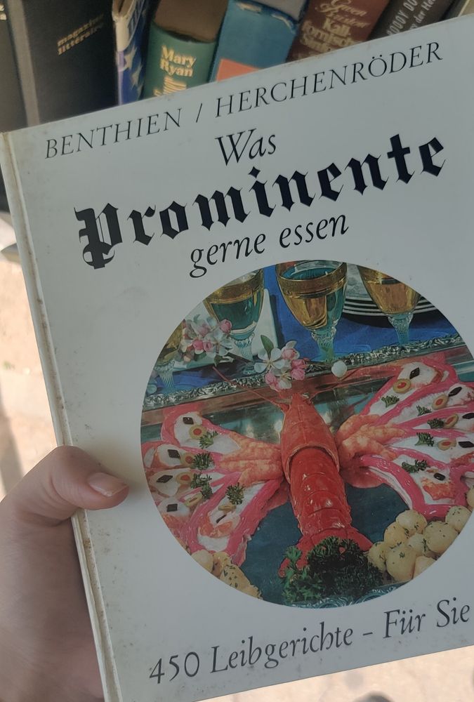 Ein altes Kochbuch mit dem Titel Was Prominente gerne essen – 450 Leibgerichte – Für Sie von Benthien und Herchenröder. Auf dem Cover ist ein kunstvoll arrangierter Hummer zu sehen, dessen Scheren und Schwanzfächer aus rosa Garnelen und weiteren Meeresfrüchten dekorativ ausgelegt sind. Der Hummer liegt auf einer Platte, umgeben von kleinen Kartoffelbällchen, Kräutern und weiteren Beilagen. Im Hintergrund stehen vergoldete Gläser und blumige Tischdekoration auf einem blauen Tuch.

