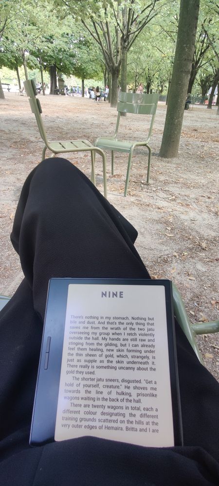 Ein eBook Reader liegt mit offenen Buch auf übereinandergeschlagenen Beinen die in einem Stuhl sitzen. Man sieht 2 metallstühle auf einer von Bäumen überdachten Fläche. Dem Jardin Luxembourg in Paris.