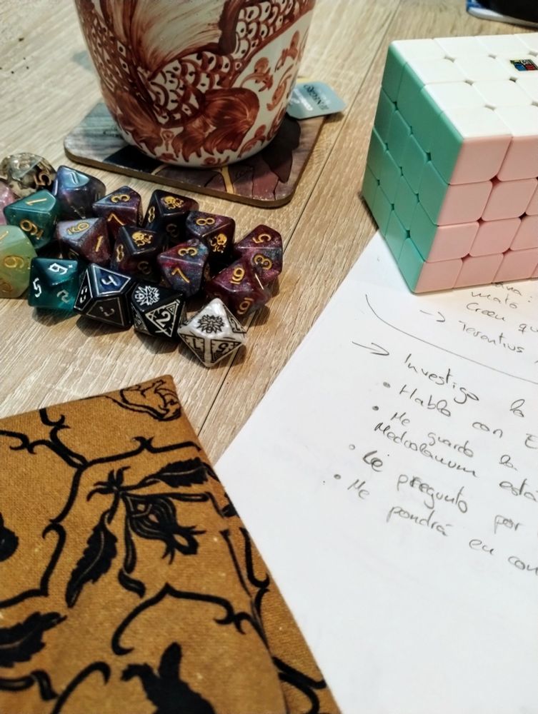 Foto de una mesa con bastantes dados d10, unos folios escritos, una bolsa de tela, un cubo de Rubik de 4x4 y una taza de té en una mesa de madera.