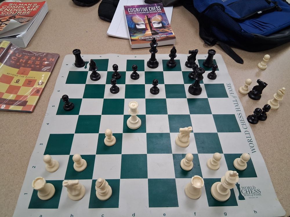 A chess board on a table with books. The position on the board has FEN:
r3kbnr/1bpp2pp/2p1p1q1/p7/3Q4/2P2N2/PP3PPP/RNB2RK1 w kq - 0 1
