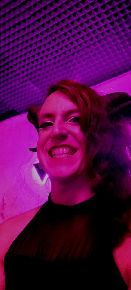 Mujer trans con cuernos de demonia enseñando los dientes bajo la luz rosada de un club nocturno. // Trans woman with demon horns, showing her teeth, glowing under the pink light of a nightclub.