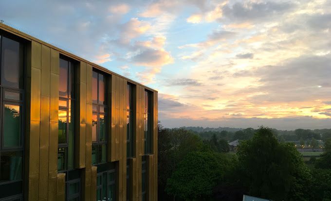 A photo of the Sir Michael Stoker Building, a research centre housed inside a golden metal cube, against the parkland of the Garscube campus as the sun rise lights up the clouds above the Campsie Fells. Image Credit: Rob Gifford.