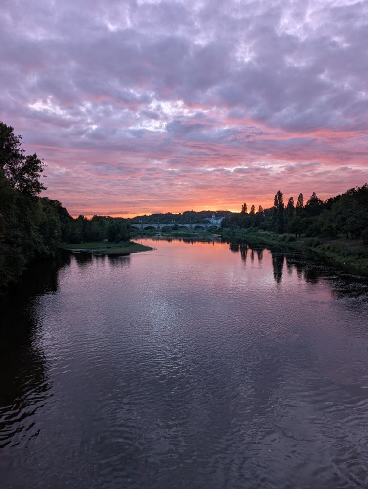 Coucher de soleil sur la Loire bordée d'arbres à Tours. Les nuages sont épais et reflètent la lumière rose - orange du soleil tandis qu'on aperçoit les arches du pont Wilson en arrière plan.