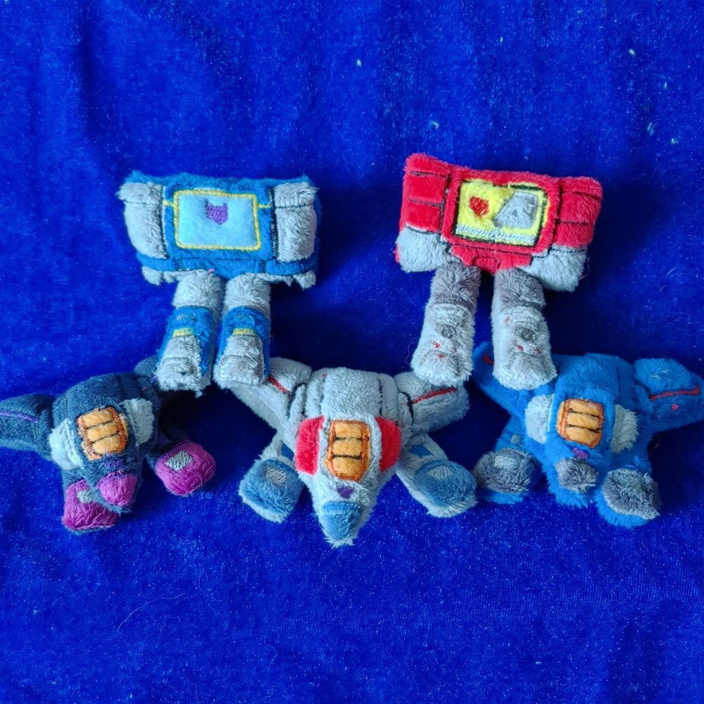 a group of five small plushies. Three of them are Thundercracker Starscream and Skywarp from Transformers; the other two are soundwave and blaster. they are all half transformed and have their legs sticking out of their alt mode forms.