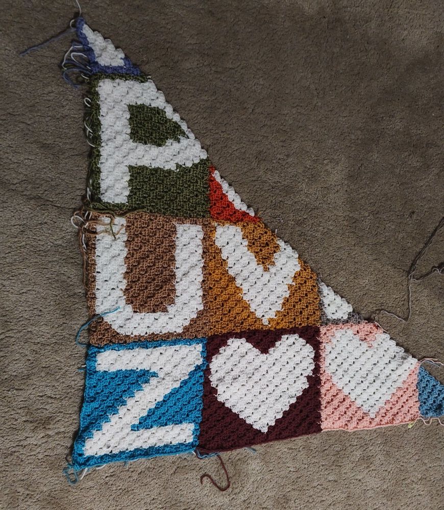 A triangular shaped blanket partially finished.  The blanket is multi-colored with letters and hearts