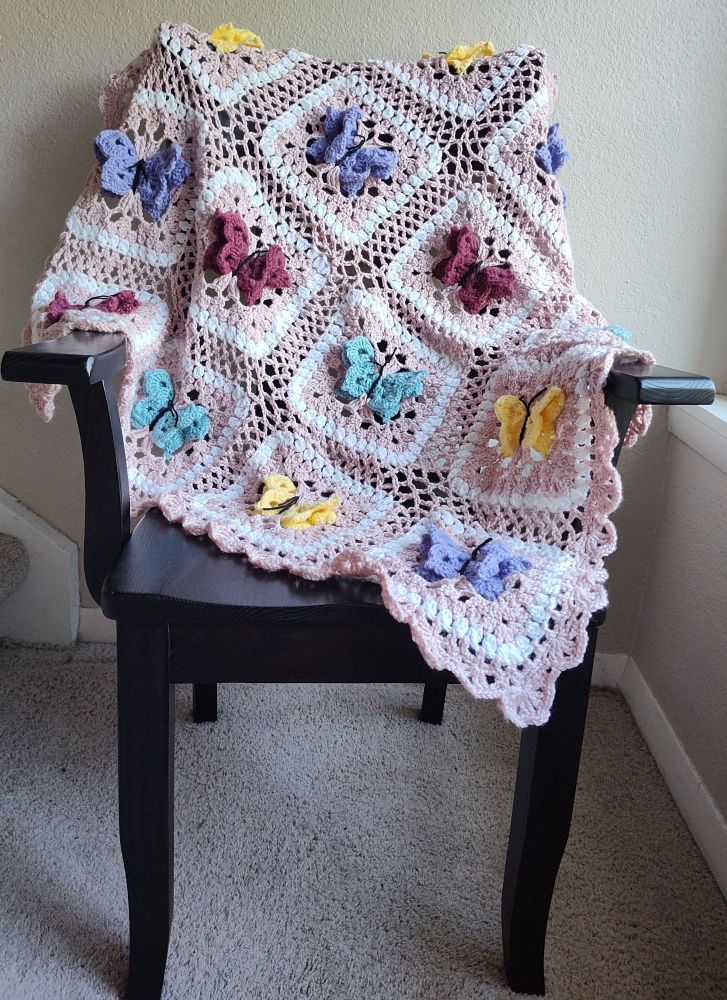 Pink baby blanket with 3D butterflies in lavender, yellow, maroon, and light blue.  Between the butterflies is a lace looking border.  The blanket is  draped on a dark brown chair, with beige wall and floor background 