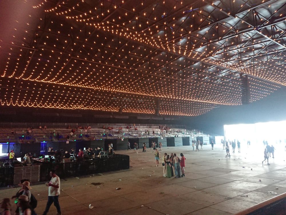Big festival tent that is still quite empty, but there are stalls on the side where people are sitting. The end of the tent is open and daylight is pouring in.