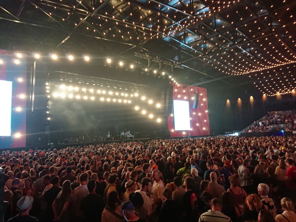 Inside a bigg festival tent with soft lights on the ceiling. The stage has been set up for the band and there are already a lot of people in the crowd.
