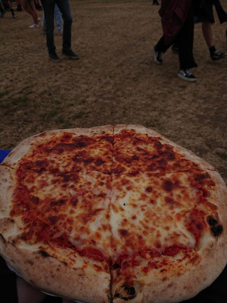 A whole pizza margarita with in the background some people on the sandy grass.