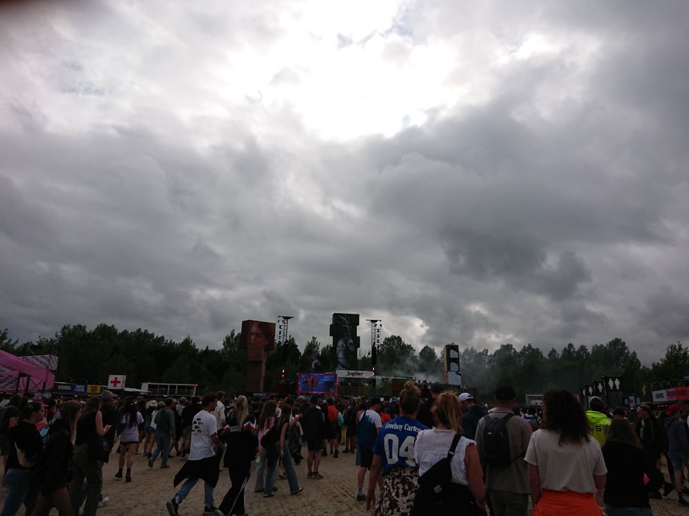Lots of people in a dance area with in the back a dj stage. The sky is full of dark clouds.
