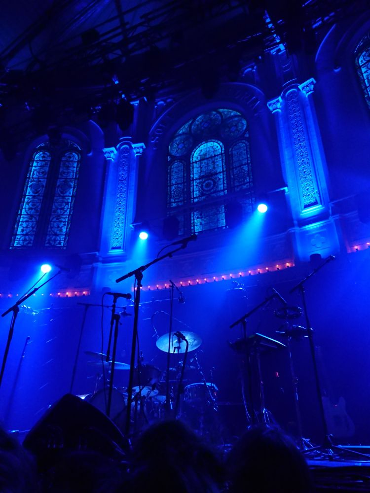 Old church turned into concert venue. The stage is lit with blue lights. Behind the old stained glass windows are visible. 