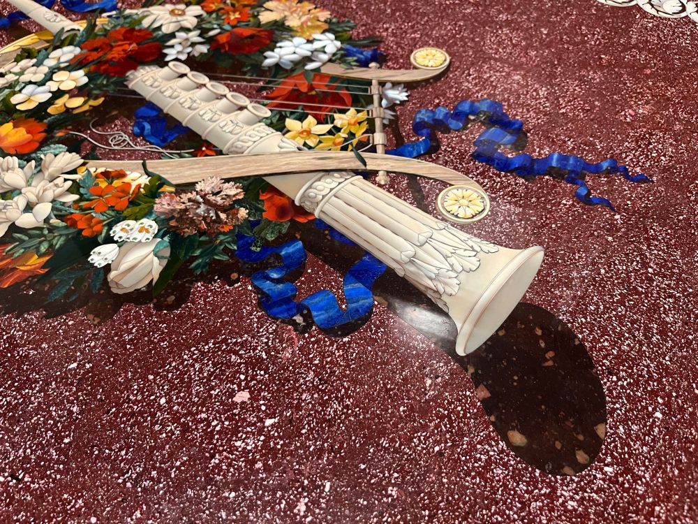 An elaborate pietre dure table top with a background of Imperial Porphry and a central emblem featuring a clarinet and a lyre surrounded by a garland of flowers and ribbons. 