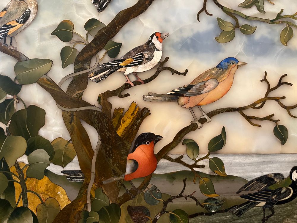 A detail of a pietre dure table top depicting a goldfinch, chaffinch and bullfinch sitting on the leafy branches of a tree. The image is created by inlaying a number of decorative hard stones. 