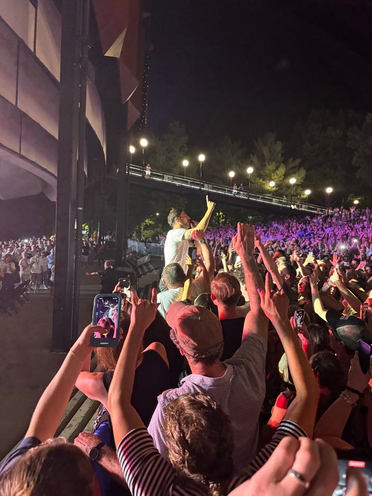 A large crowd at a Mumford & Sons concert surrounds the lead singer, who stands in the middle of the audience with one arm raised as fans reach toward him. Many people are capturing the moment with their phones. The night sky and stage lights create a glowing, electric atmosphere.
