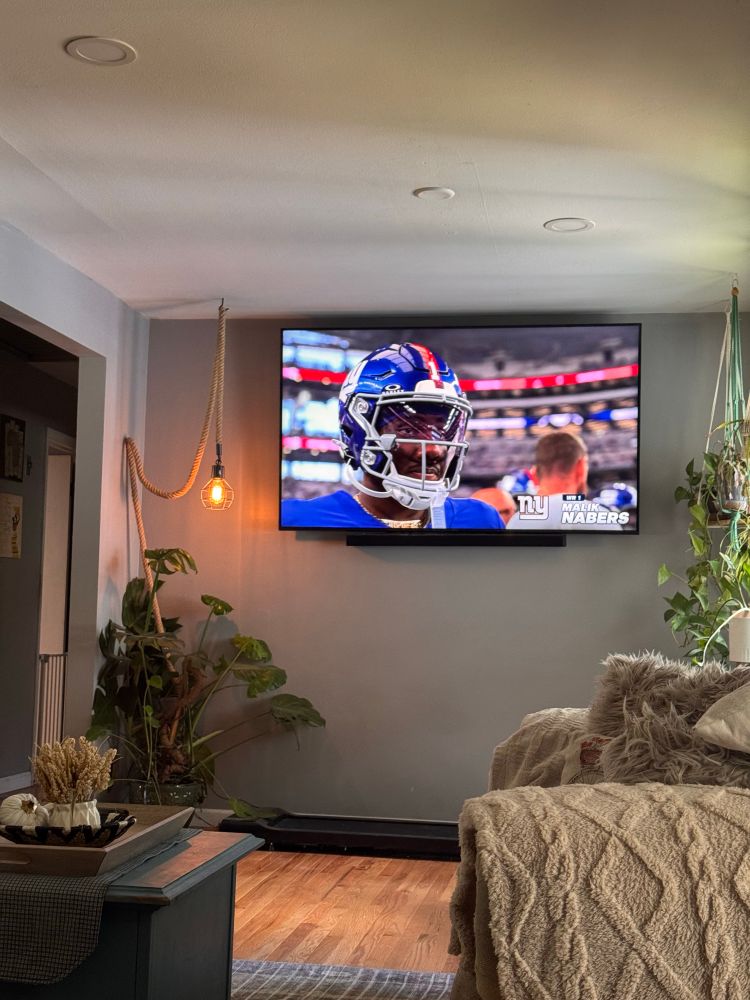 Living room with cozy neutral decor, plants, and a big screen TV showing a New York Giants player in uniform during the game.