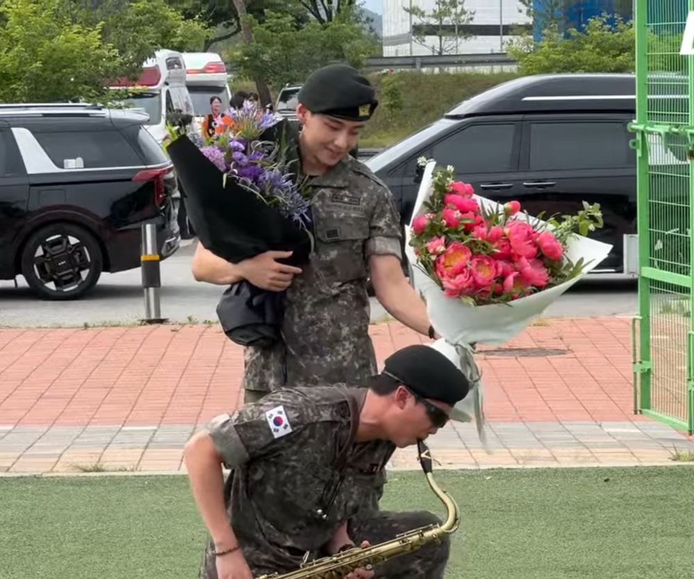 kim namjoon down on one knee enthusiastically playing the saxophone in front of kim taehyung holding a bouquet of flowers for himself, and holding another out to namjoon before him