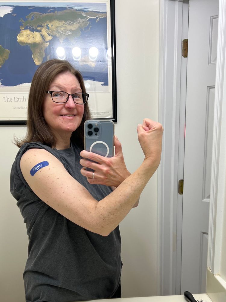 A smiling woman with dark hair and glasses is flexing her right arm which has a Kroger band aid on it. 