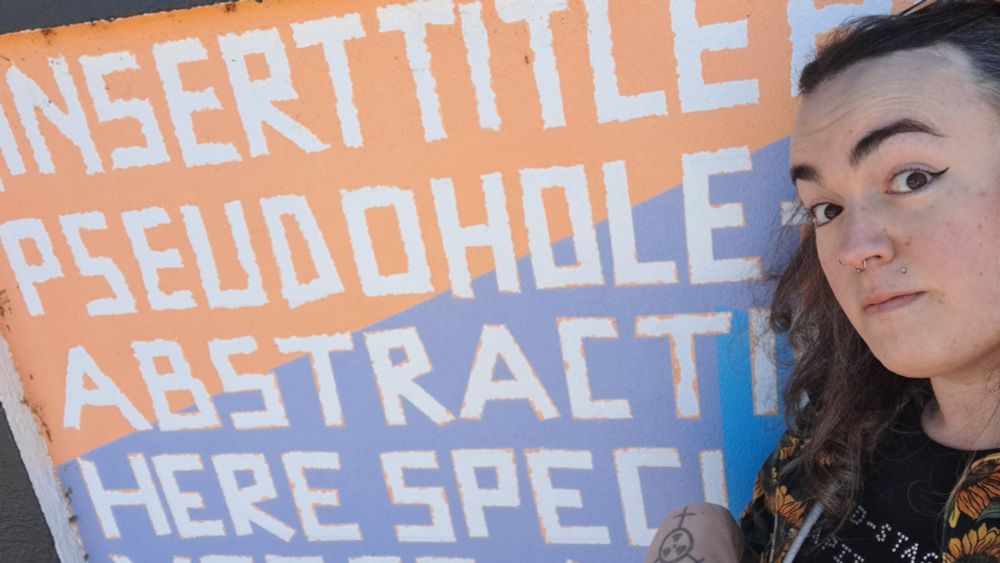 A woman standing in front of a mural, wide-eyed, the mural has a series of words and phrases written across it including the word "Pseudohole"