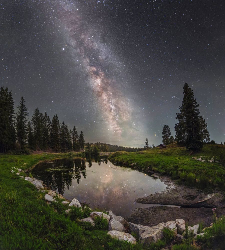 Summer Milky Way over a small lake reflecting the Milky Way. Lake has rocks piled up around with tress in the background. Southern BC