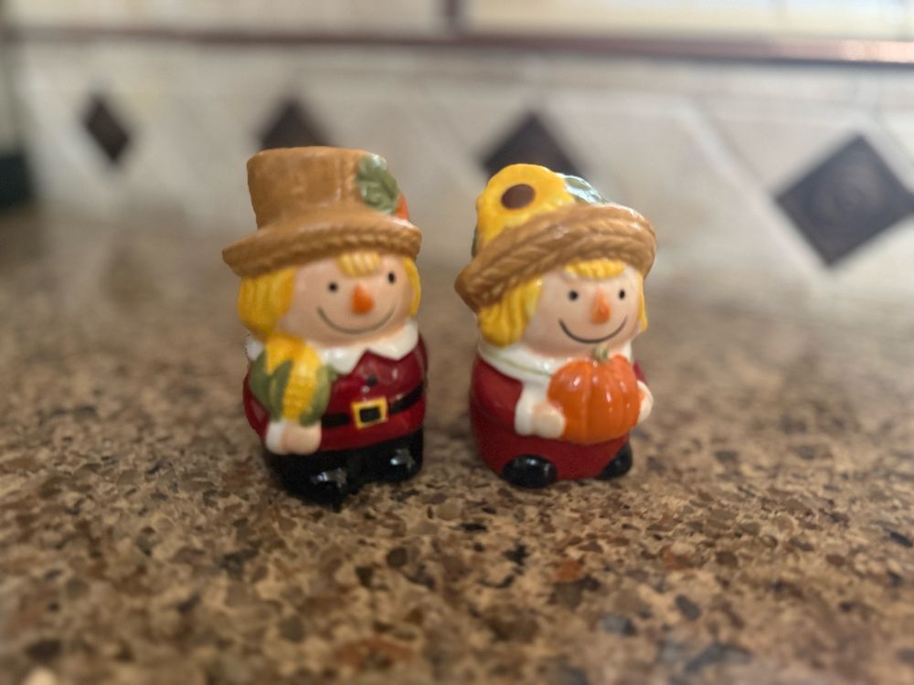 Straight ahead photos of a salt and pepper shaker designed as two farmers. The one on the left is a boy holding an ear of corn. The girl on the right holds a pumpkin in two hands and has a sunflower on her hat. They are both smiling. 