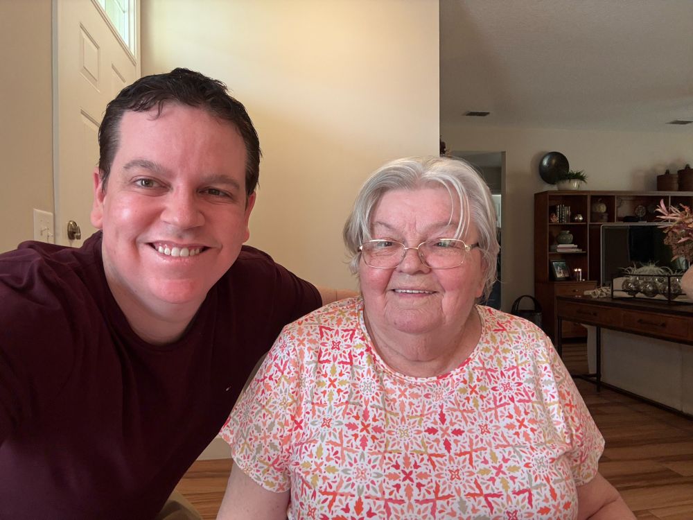 A son on the left with his mom at a table. She is wearing a colorful patterned shirt of red, orange, green and yellow. 