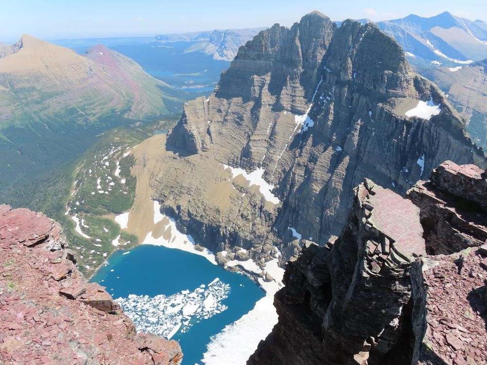 View of reddish rocks looking down at blue lake with white ice and serrated tall rocky ridge on one end. 