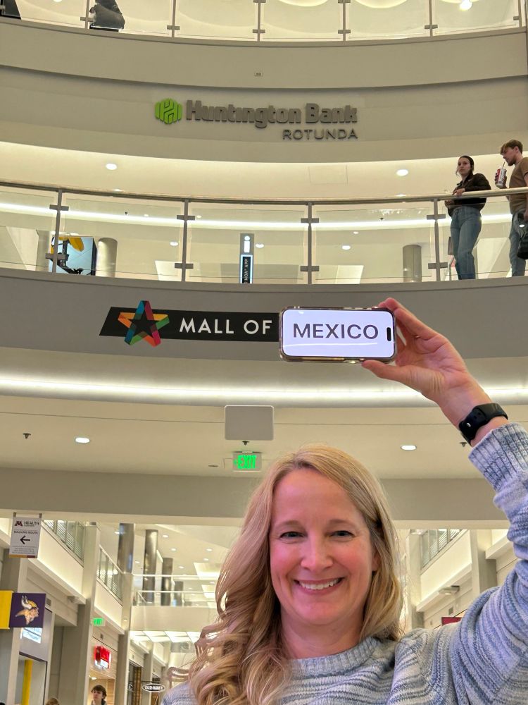 A woman stand in front of the sign for Mall of America and holds the word Mexico in front of the word America. 