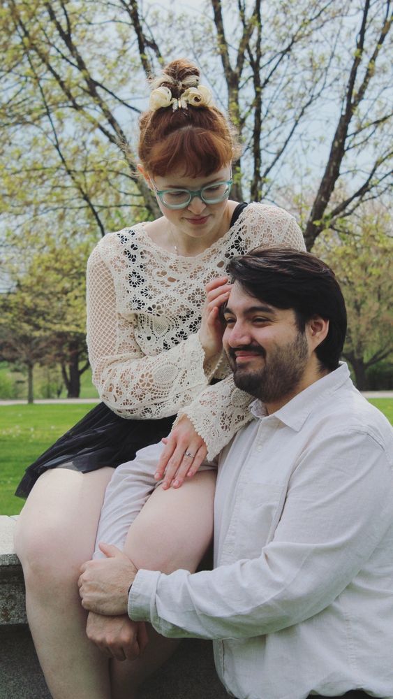 Me and my fiancé posing next to each other in front of trees. I am sitting on a concrete slab, and I have my right hand resting towards his face.