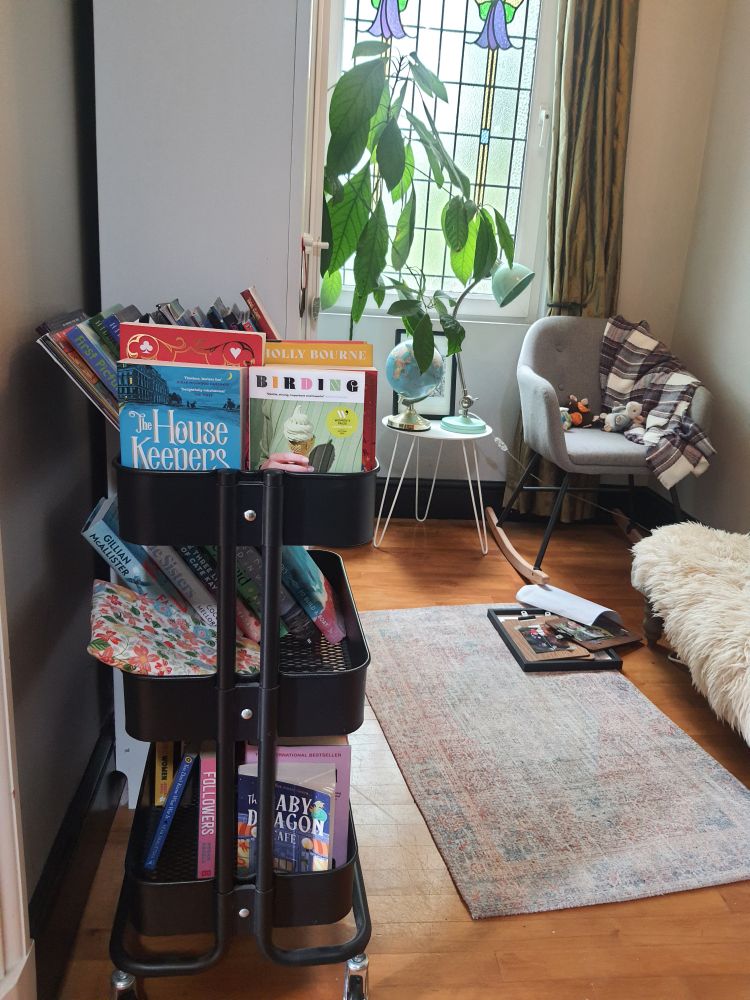 Left: A black trolley of books with big white bookcase behind. 'Birding' is on the trolley. Window and Avocado tree in the background. Chair and mini table next to it also by the window. Chair has a woolly blanket and some kids' knitted toys on it. Globe and picture in frame and lamp on table. Wooden floor with a rug on it. Some photos of the kids on the floor. Right of picture is footstool with a sheepskin rug on it.