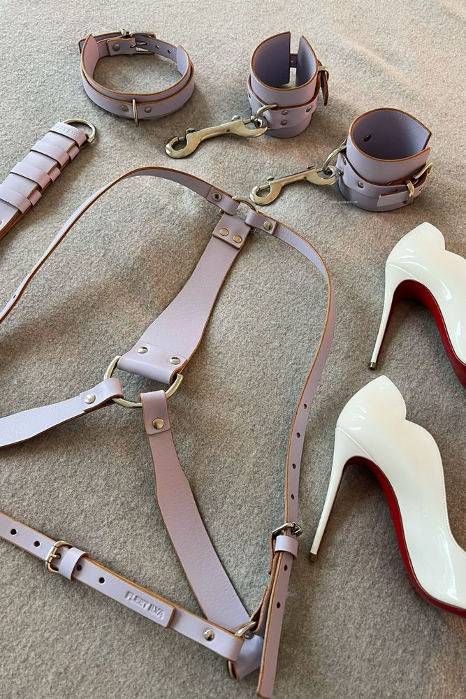 A lilac Fleet Ilya leather harness and cuffs set with flogger, and white Louboutin high heels, laid out on a bed