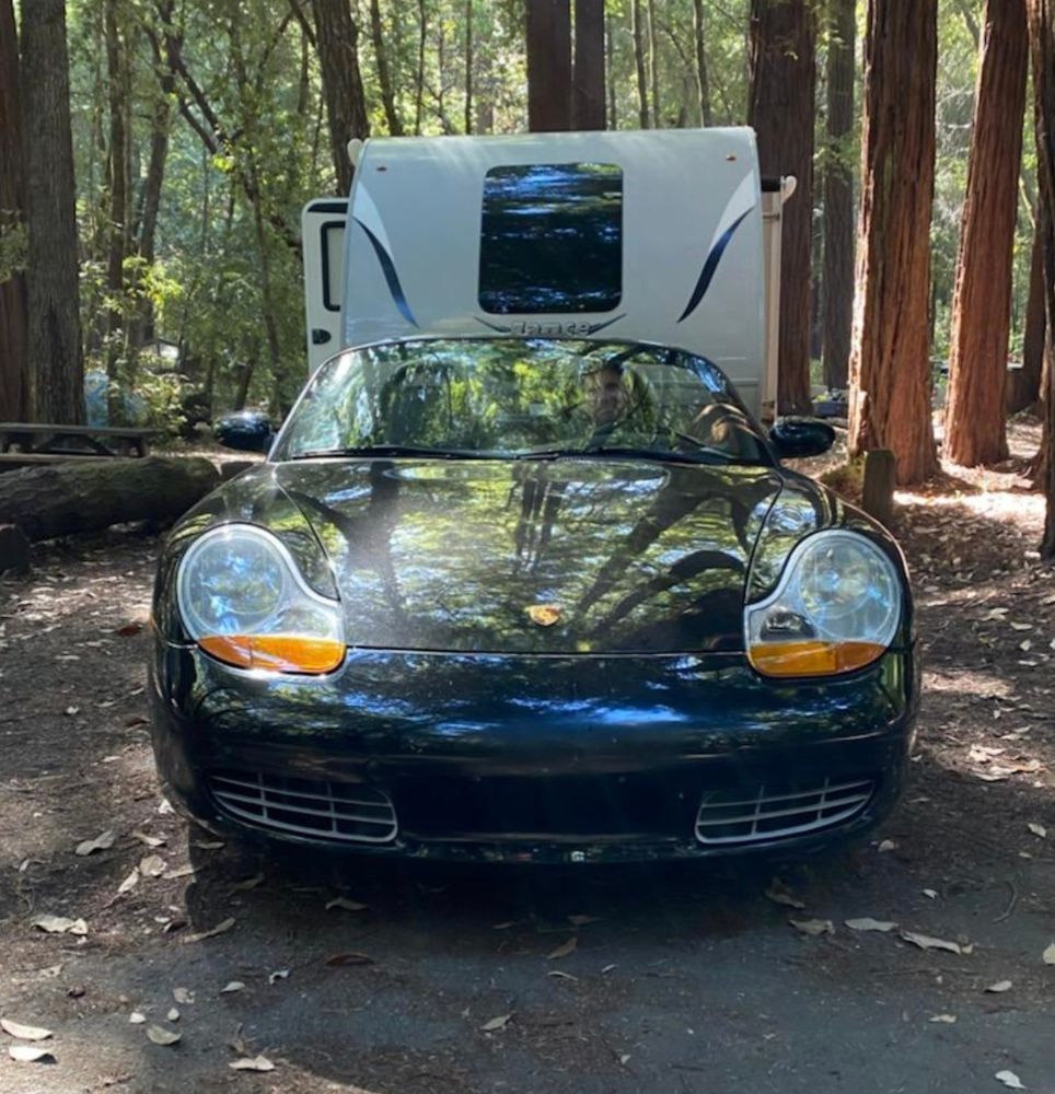Black 1998 Porsche Boxster parked in front of a Lance camping trailer in a redwood forest. 
