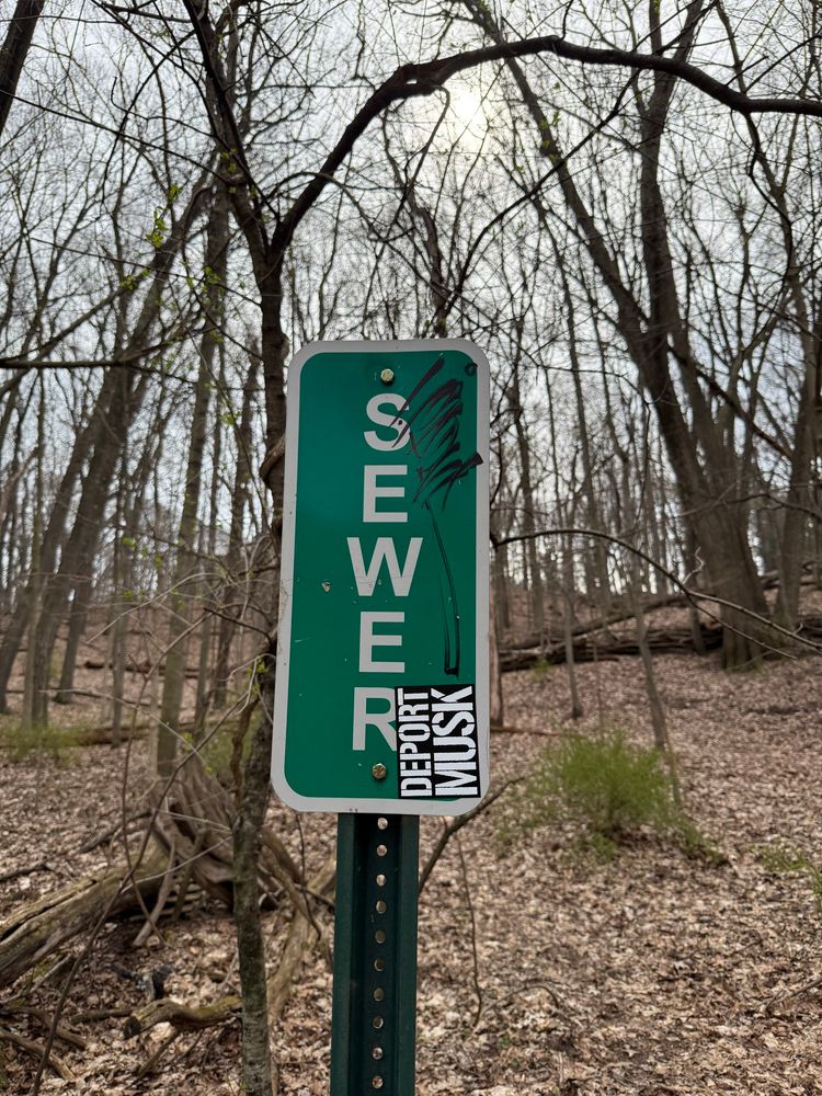 A sewer sign with a sticker that says deport most attached to it. 