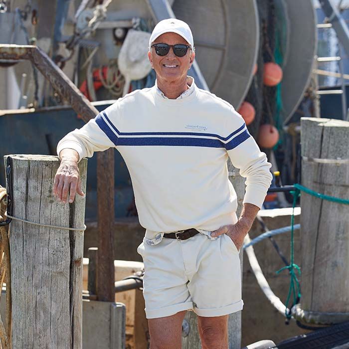 older man wearing pale khaki shorts (ugh) with a belt and a light white sweater with a blue stipe, sunglasses and a white baseball cap (ugh) standing on a dock with a fishing boat in the background