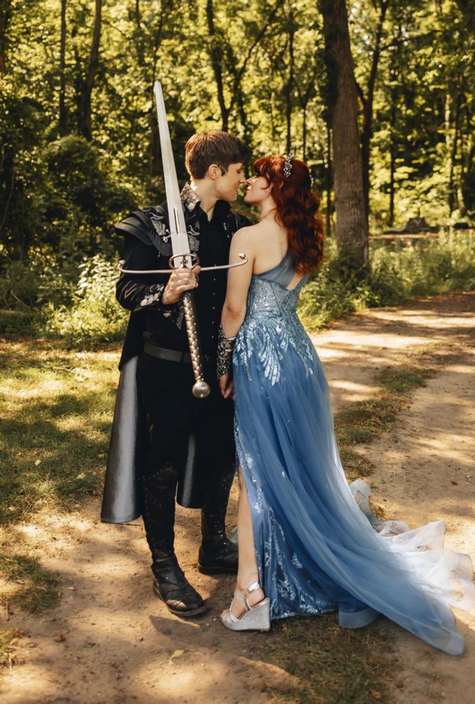 photo of Claire and husband Drew on their wedding day: him in black-and-silver jacket with cape and pauldrons and holding a greatsword, her in a shimmery periwinkle-blue dress and crown