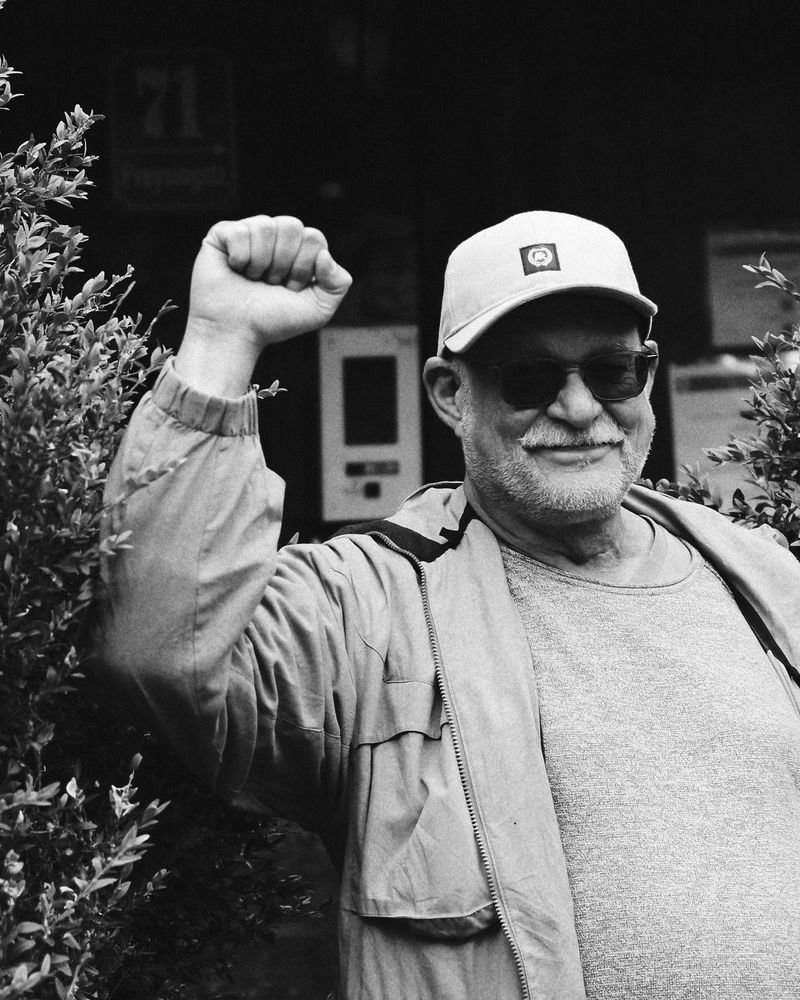 Schwarzweißes Portrait eines lächelnden Jörg mit Bart, Sonnenbrille und Baseballkappe, der die Faust in einer Geste der Solidarität und des Kampfgeistes erhebt. Er trägt eine helle Jacke und ein T-Shirt. Im Hintergrund sind unscharfe Blätter einer Pflanze zu sehen. Das Bild strahlt sowohl Entschlossenheit als auch Wärme aus.