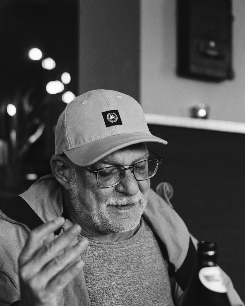Das schwarz-weiße Porträtfoto zeigt Jörg im Profil. Er trägt eine Baseballkappe mit einem kleinen Logo-Patch, eine Brille und hat einen grauen Bart. Der Mann lächelt leicht und trägt einen Pullover mit dünner Regenjacke. Die Aufnahme hat eine künstlerische Qualität mit schöner Bokeh-Beleuchtung im Hintergrund und zeigt den Mann in einem nachdenklichen, entspannten Moment.
