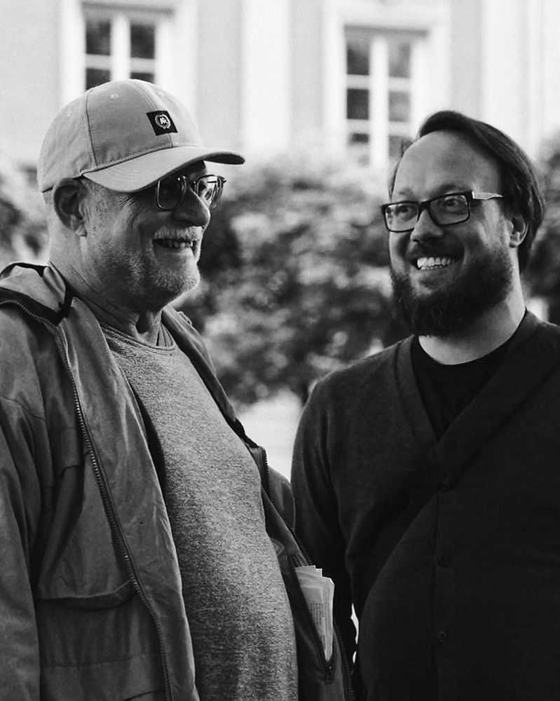 Ein Schwarzweißfoto von zwei lächelnden Männern. Der ältere Mann, Jörg, auf der linken Seite trägt eine Baseball-Kappe, eine Jacke und eine Brille. Der jüngere Mann, Co-Gründer der Stiftung Konstantin, auf der rechten Seite hat einen Bart und trägt ebenfalls eine Brille. Beide stehen vor einem hellen Gebäude mit Fenstern im Hintergrund und strahlen in die Kamera. Das Bild vermittelt eine warme, freundschaftliche Atmosphäre.