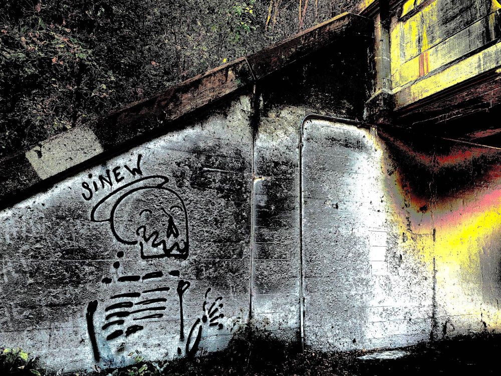 A photo of graffiti and a bridge. The graffiti is of a happy skeleton with a caption saying "sinew". Above this is some trees and bushes on a slope. The bridge curves up and the colors of the overpass are distorted and manipulated into saturation.
