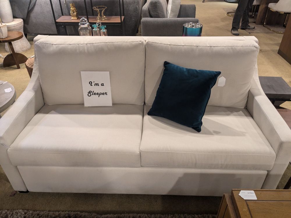 Photo of a white sofa in a furniture store that has a sign on it that says "I'm a sleeper."