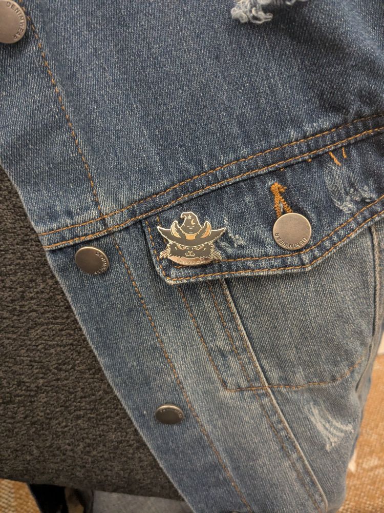 A denim vest draped over the back of a chair with an enamel pin of a cat wearing a witch hat on the pocket.