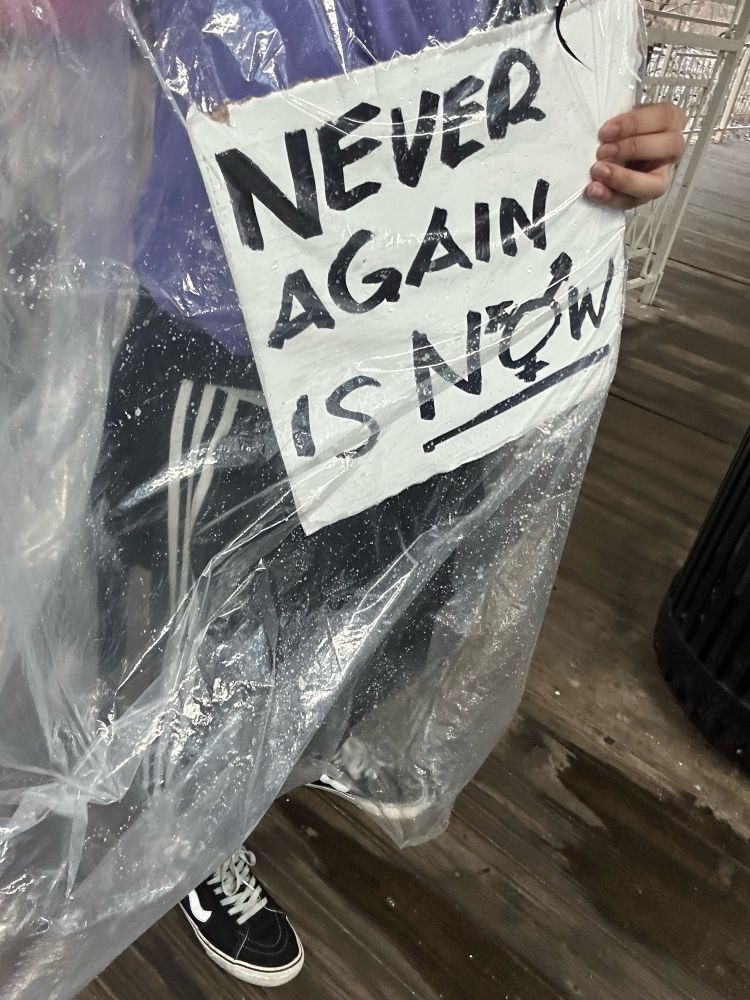 Sopping fucking wet creature holds signage saying “never again is now”