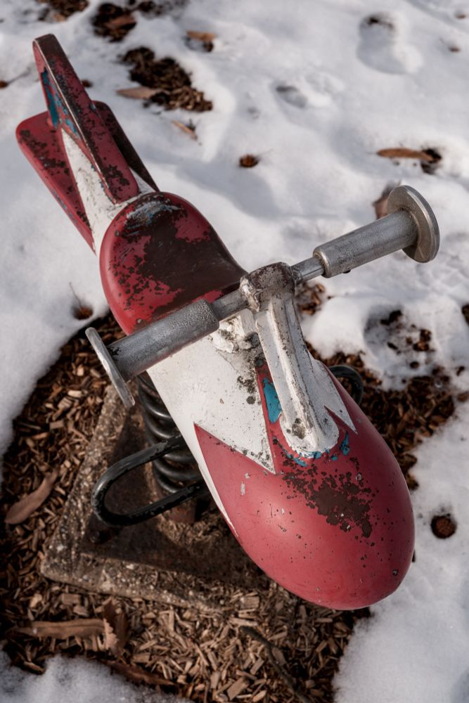 Photograph of a red and white space age rocket-shaped spring ride on a partially snow covered playground.  The paint is faded in spots, and there are a few flecks of blue.